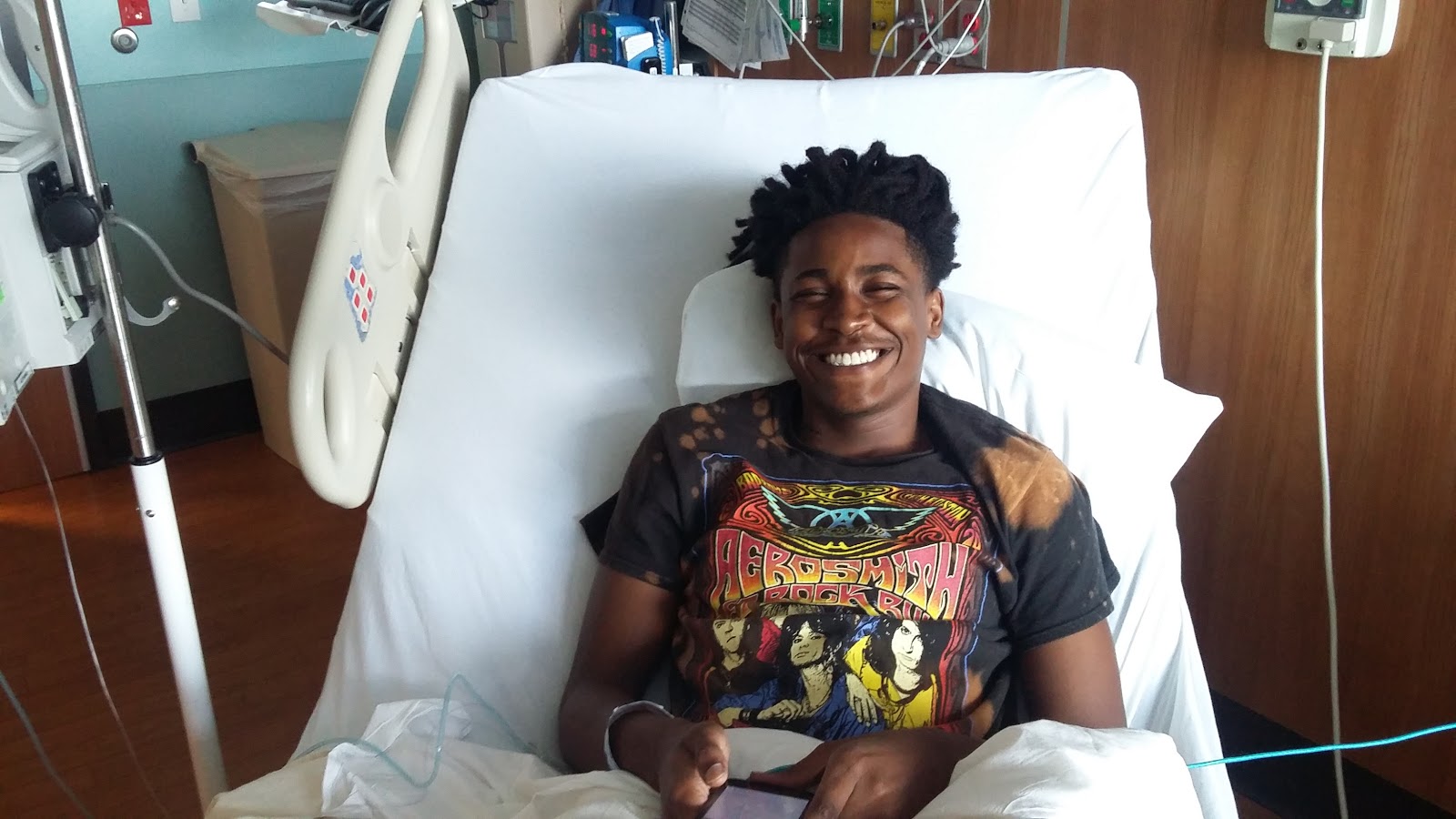 Qevonte laughing in his hospital bed wearing an Aerosmith t-shirt, surrounded by pillows and cards from supporters.