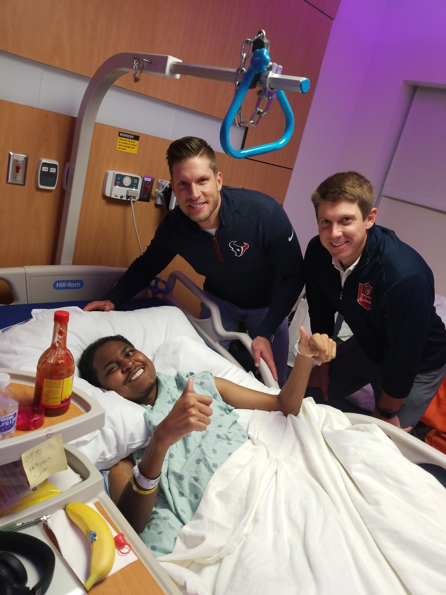 Qevonte giving a proud thumbs up from his hospital bed while Houston Texans football players visit him at his bedside.