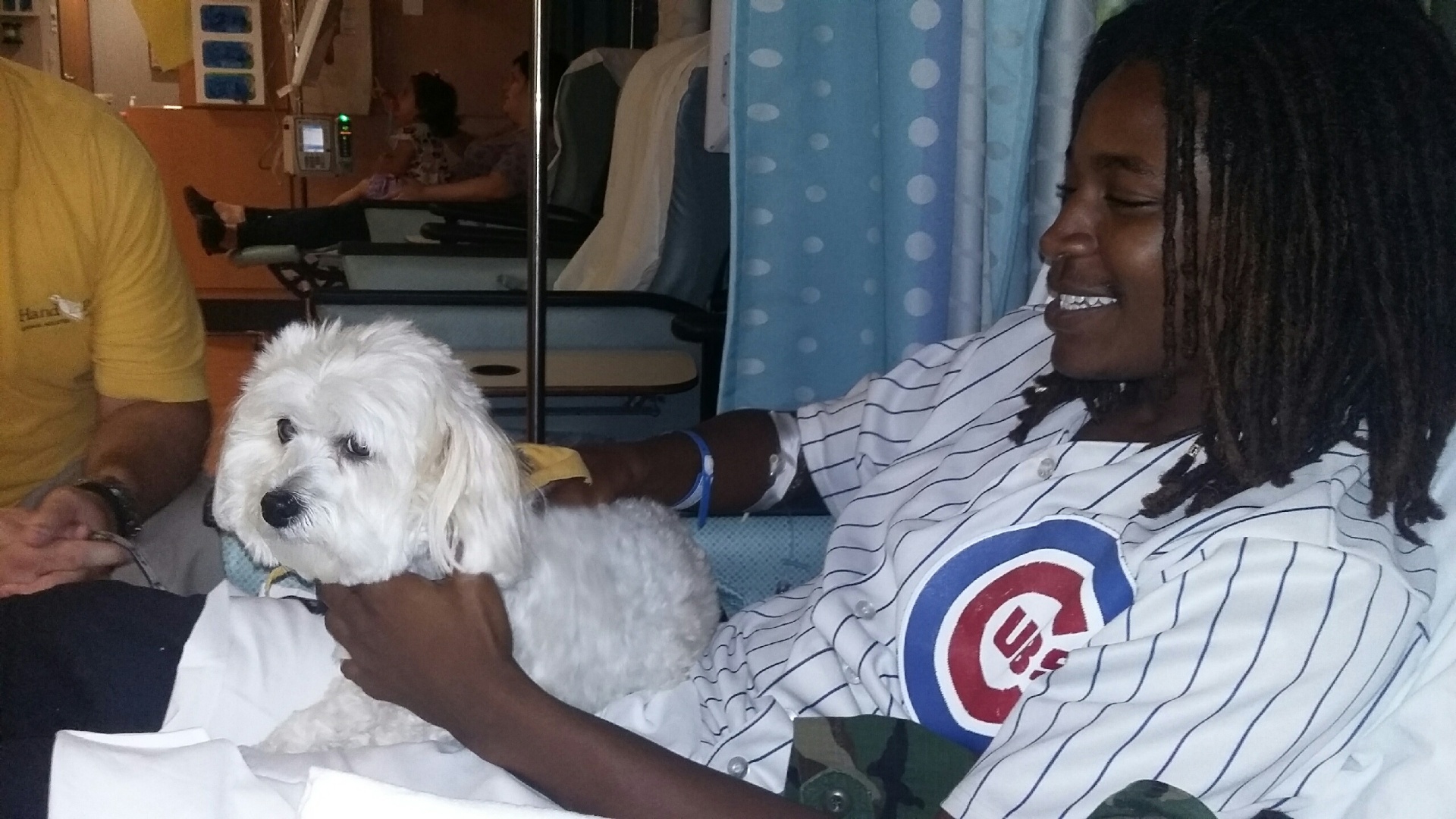 Qevonte smiling softly as he reaches out to pet a golden therapy dog resting beside him in the hospital.
