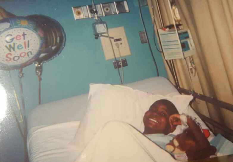 Qevonte sitting up in his hospital bed, beaming a wide smile next to a bright 'Get Well Soon' balloon left by visitors.